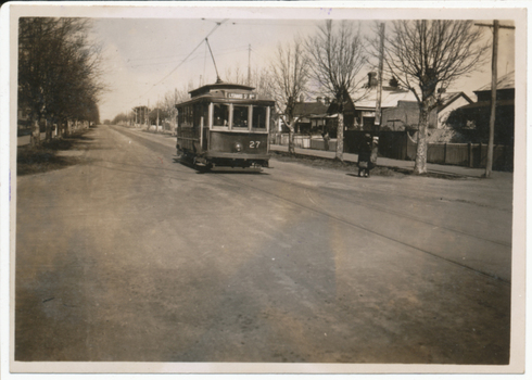 Vintage tram numbered 27 traveling down a tree-lined street in a residential area.  A lady is walking across to the footpath after alighting from the tram.