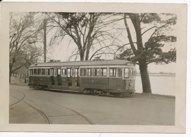 A vintage electric tramcar with multiple windows, parked on a track next to a body of water and several bare trees.
