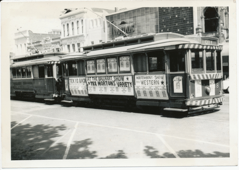 Two vintage trams are parked on a city street, covered in advertisements for "Tex is Back at the Ballarat Show" and "Hootenanny Show and Western," featuring "Tex Morton's Variety."