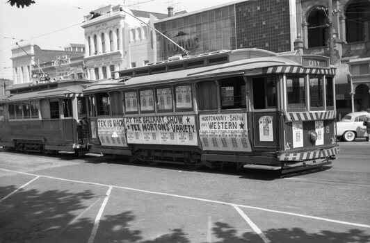 Two vintage trams are parked on a city street, covered in advertisements for "Tex is Back at the Ballarat Show" and "Hootenanny Show and Western," featuring "Tex Morton's Variety."