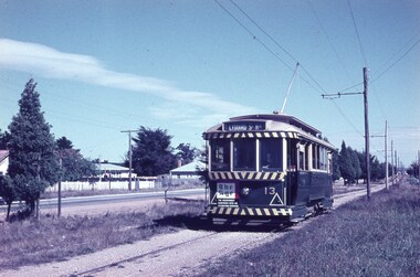 A vintage dark green tram with a striped roof and destination board marked "Lydiard St Nth" travels on tracks beside a road lined with trees and houses.