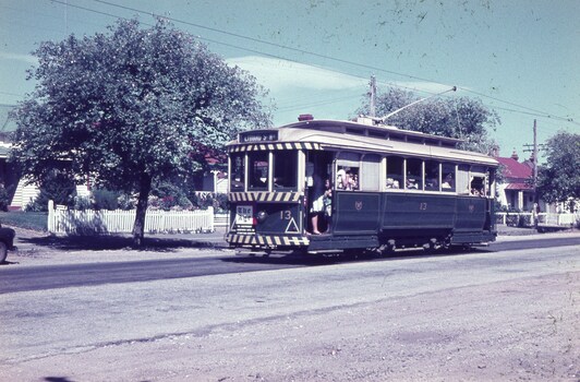Vintage green tram with striped front, carrying passengers, traveling on a paved street in a suburban area with houses and trees.