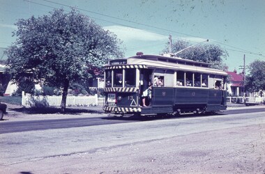 Vintage green tram with striped front, carrying passengers, traveling on a paved street in a suburban area with houses and trees.