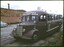 A maroon vintage bus with "SQUIRES BALLARAT-AVOCA" written in white text on its front, parked on a dirt road next to a brick building with a "Gold" sign and a small white car.