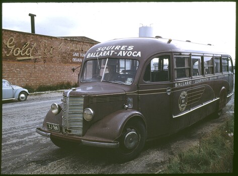 A maroon vintage bus with "SQUIRES BALLARAT-AVOCA" written in white text on its front, parked on a dirt road next to a brick building with a "Gold" sign and a small white car.