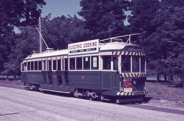 A vintage dark green and cream tram with "ELECTRIC COOKING" signage on top, parked on a paved road lined with trees.