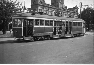 A vintage, dark-colored tram with open doors and passengers visible inside travels along a paved street in front of an ornate, historic building.