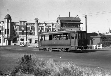 A vintage non passenger tram stands on tracks in front of a grand brick building with ornate architectural details and a domed tower, next to a wooden signal box.