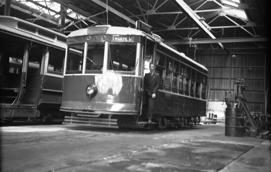 A man in a dark suit stands in the open doorway of a vintage tram labeled "THORPE ST" inside a cavernous industrial workshop or tram depot.