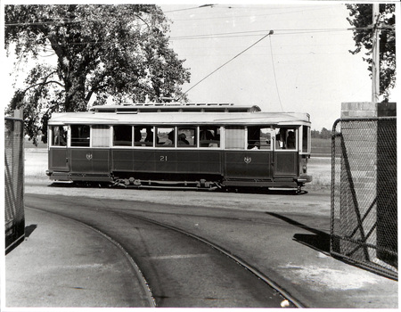 Vintage tramcar numbered 21 with passengers visible inside, traveling on tracks on a sunny day, with a fence in the foreground.
