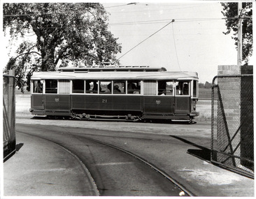 Vintage tramcar numbered 21 with passengers visible inside, traveling on tracks on a sunny day, with a fence in the foreground.