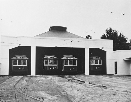 Four vintage trams are parked inside an open-sided depot with a rounded roof, under overhead electrical wires.