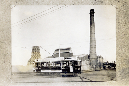 A black and white photograph shows a vintage tram with the words "BALLARAT TRAMWAYS" and "FOLLOW THE CROWD TO HARRYS SALE" printed on its side and roof. The tram has large windows displaying advertisements for "COLLESEUM STAR MOVIES," and appears to be traveling down a street with industrial buildings and a tall smokestack in the background.