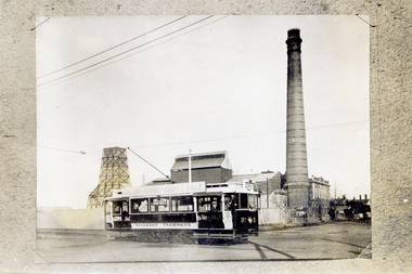 A black and white photograph shows a vintage tram with the words "BALLARAT TRAMWAYS" and "FOLLOW THE CROWD TO HARRYS SALE" printed on its side and roof. The tram has large windows displaying advertisements for "COLLESEUM STAR MOVIES," and appears to be traveling down a street with industrial buildings and a tall smokestack in the background.