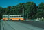 An articulated bus with orange and white livery travels on a street with tram tracks in front of a dense line of green trees under a clear blue sky.