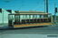 A vintage wooden tram car, labeled "M & MTB," with a man in a suit sitting at the controls, moves along tram tracks in an urban setting with overhead power lines and industrial buildings.
