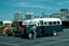 A row of vintage buses are parked in an outdoor lot with city buildings in the background.