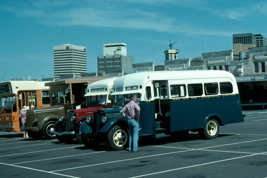 A row of vintage buses are parked in an outdoor lot with city buildings in the background.