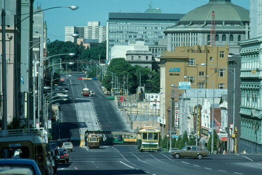 A wide, sunny street slopes downhill, lined with buildings on both sides. Several vintage trams, some yellow and green and one red, travel up and down the street.