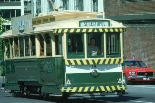 A green and cream vintage tram with yellow and black striped trim travels along a street.