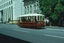 A red and white open-sided tram numbered 17 travels along a city street with ornate buildings and trees in the background.