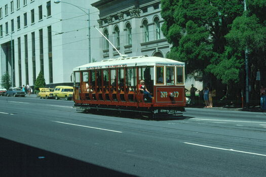 A red and white open-sided tram numbered 17 travels along a city street with ornate buildings and trees in the background.