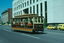 Vintage wooden tram with open sides and passengers, moving on city street past yellow cars and buildings.