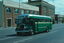A green and silver vintage bus with "SPENCER ST" and "VICTORIA MARKET" displayed on its route indicator drives along an asphalt road past brick buildings under a cloudy sky.