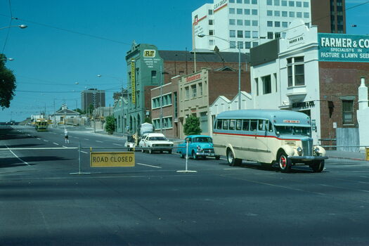 A vintage cream and light blue bus with red trim drives past a "ROAD CLOSED" sign on a street lined with buildings and a tram.