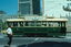 A green and cream vintage tram with the number 14 on its side crosses a street in front of modern buildings, with a person walking across the street in the foreground.