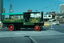 A green Ransome Steam Wagon with a canopy over the back, with "RANSOMES, SIMS & JEFFERIES LTD IPSWICH" printed on its side, drives through a city intersection.