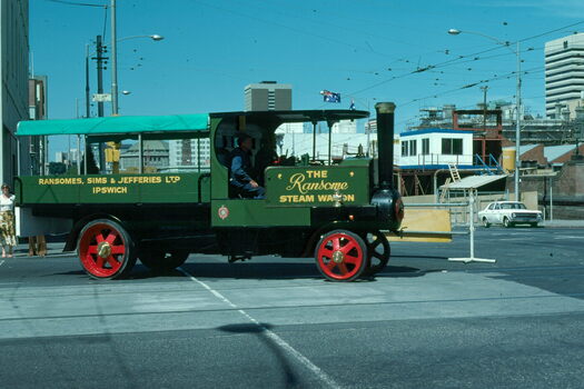 A green Ransome Steam Wagon with a canopy over the back, with "RANSOMES, SIMS & JEFFERIES LTD IPSWICH" printed on its side, drives through a city intersection.
