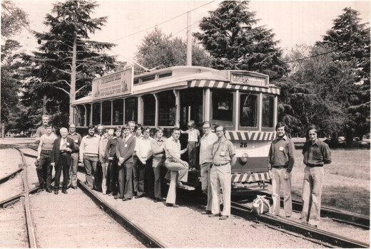A group of men and one child pose for a photo in front of a vintage tram. The tram has a sign that reads "GARDENS" on the front.