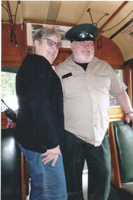 A woman and a man in a tram car, the man wearing a uniform cap and shirt with sunglasses perched on the cap.