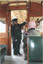 A uniformed conductor with a cap and sunglasses stands in the doorway of a vintage wooden tram car, holding onto the door frame.