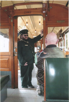 A uniformed conductor with a cap and sunglasses stands in the doorway of a vintage wooden tram car, holding onto the door frame.