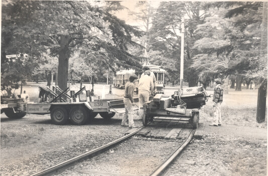 A trolley sits on tracks with people working on a trailer carrying heavy equipment next to the tracks, with tramcars and trees in the background.