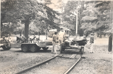A trolley sits on tracks with people working on a trailer carrying heavy equipment next to the tracks, with tramcars and trees in the background.