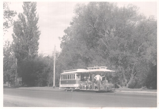 A vintage cable car, numbered 171, and a motorised grip car carries passengers along a road lined with trees. The open-air cable car has a white roof and several people are visible, some standing and some seated.