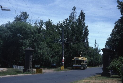 A vintage yellow and green tram travels down a tree-lined road past stone pillars. A street lamp is visible in the upper left corner against a partly cloudy sky.