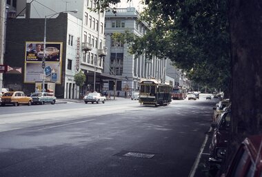 A vintage tram moves down a city street lined with trees, cars, and buildings, including the Phoenix Hotel.