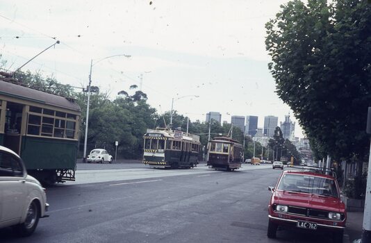 A vintage tramway scene with multiple vintage trams and cars on a city street with tall buildings in the background.