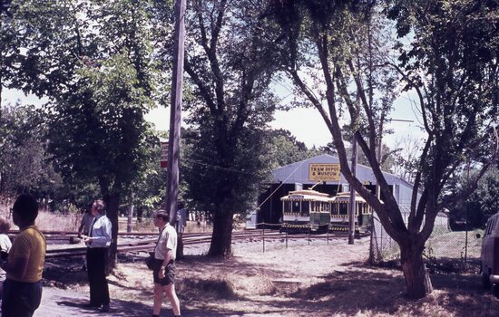 Two vintage trams are parked in an open-sided depot and museum building, with a complex arrangement of tram tracks in the foreground. A sign above the tram entrances reads "TRAM DEPOT & MUSEUM."