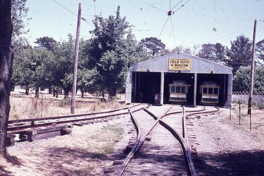 Two vintage trams are parked in an open-sided depot and museum building, with a complex arrangement of tram tracks in the foreground. A sign above the tram entrances reads "TRAM DEPOT & MUSEUM."