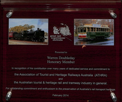 Plaque with "Presented to Warren Doubleday Honorary Member" text and two inset images: one of a black steam locomotive pulling red carriages, and another of a green and yellow vintage tram, along with a "Great Rail Experiences Australia" logo.