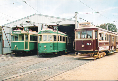 Three vintage trams, two green with cream accents and one maroon with cream accents, parked in front of a corrugated metal tram shed.