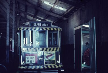 A vintage yellow and black striped tram labeled "DEPOT VIA RIPON ST" is parked inside a dimly lit corrugated metal shed, featuring advertisements for "The Age" newspaper and "Mutual Homes."