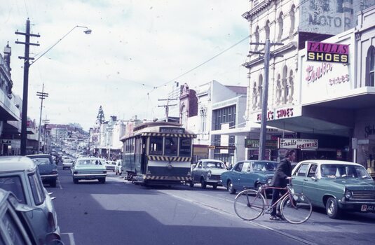 A vintage green and cream tram travels down a busy street lined with shops, including "Faulls Shoes," with cars and a bicycle on the road under an overcast sky.