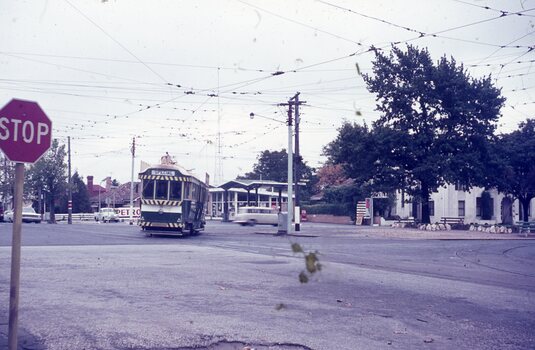 A green and white tram marked "SPECIAL" travels on a street with overhead power lines, passing a petrol station and buildings, with a red stop sign in the foreground under an overcast sky.