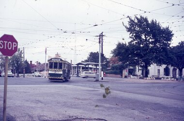 A green and white tram marked "SPECIAL" travels on a street with overhead power lines, passing a petrol station and buildings, with a red stop sign in the foreground under an overcast sky.